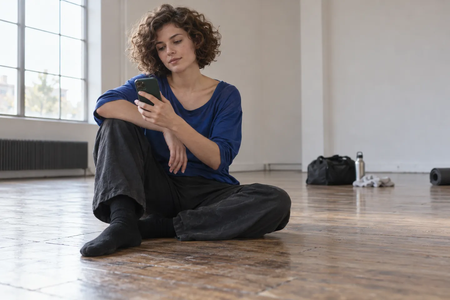 Young adult with a short natural curly bob considering a phone while seated on the floor of a quiet rehearsal studio