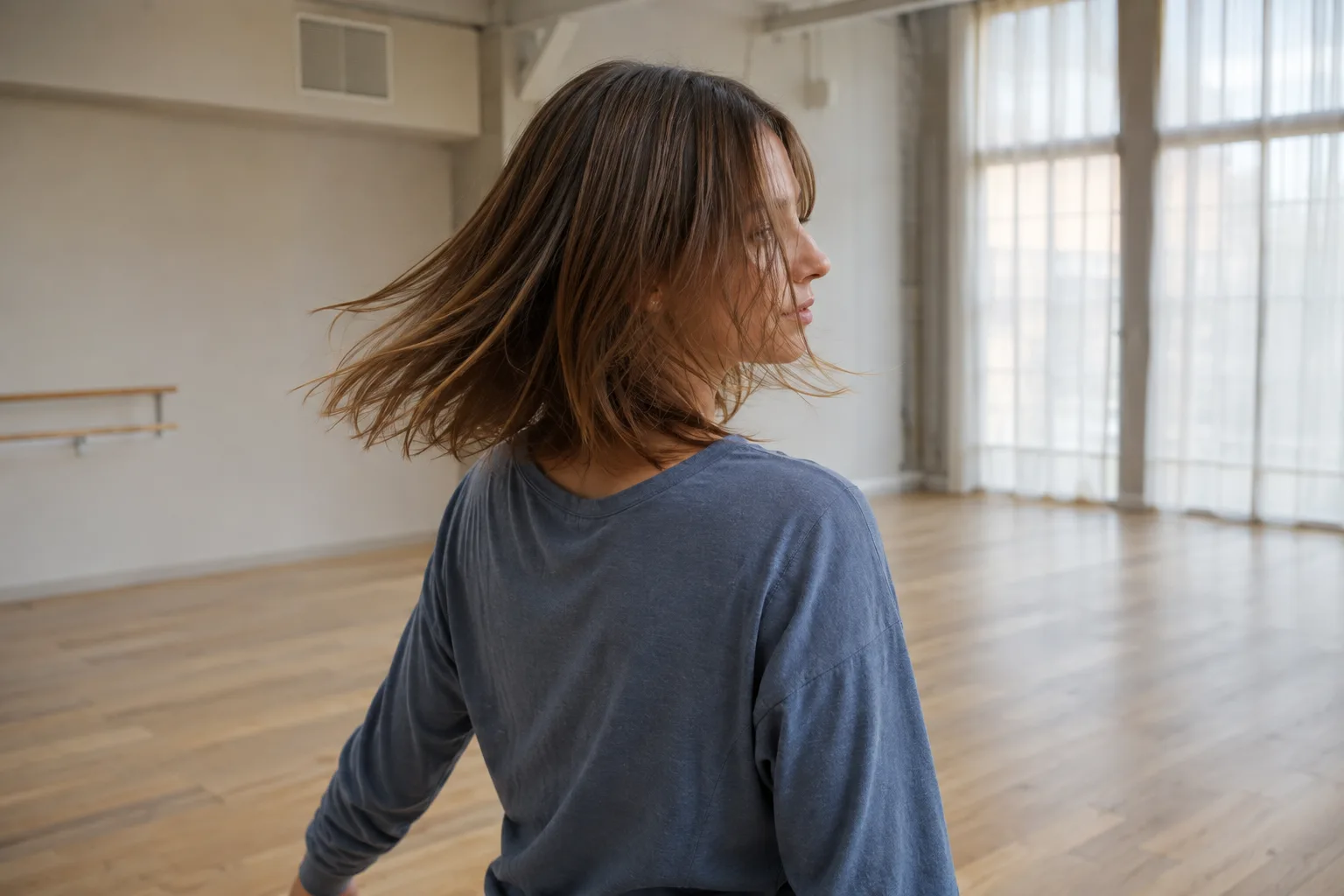 Young woman with fine shoulder-length brown hair turning gently in a bright dance studio to check the moving haircut outline