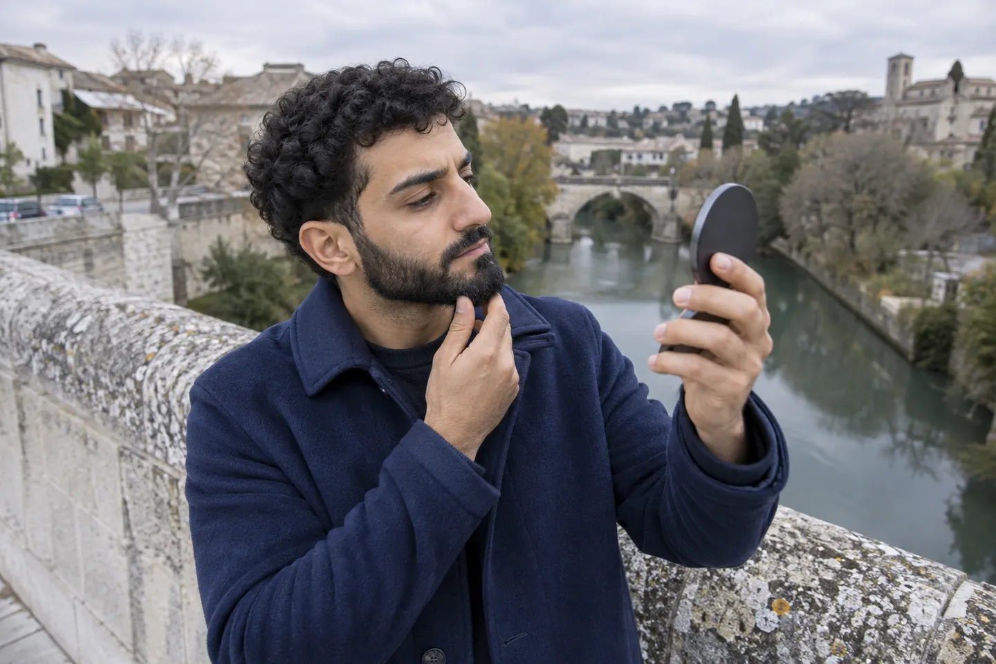 Young man with short black curls and a full beard studying his outline in a hand mirror on a stone footbridge