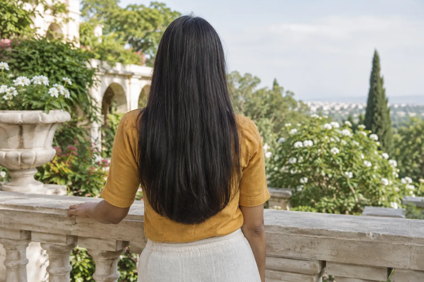 Young woman with long black hair and a softly rounded lower outline leaning on a pale stone terrace balustrade, seen from behind