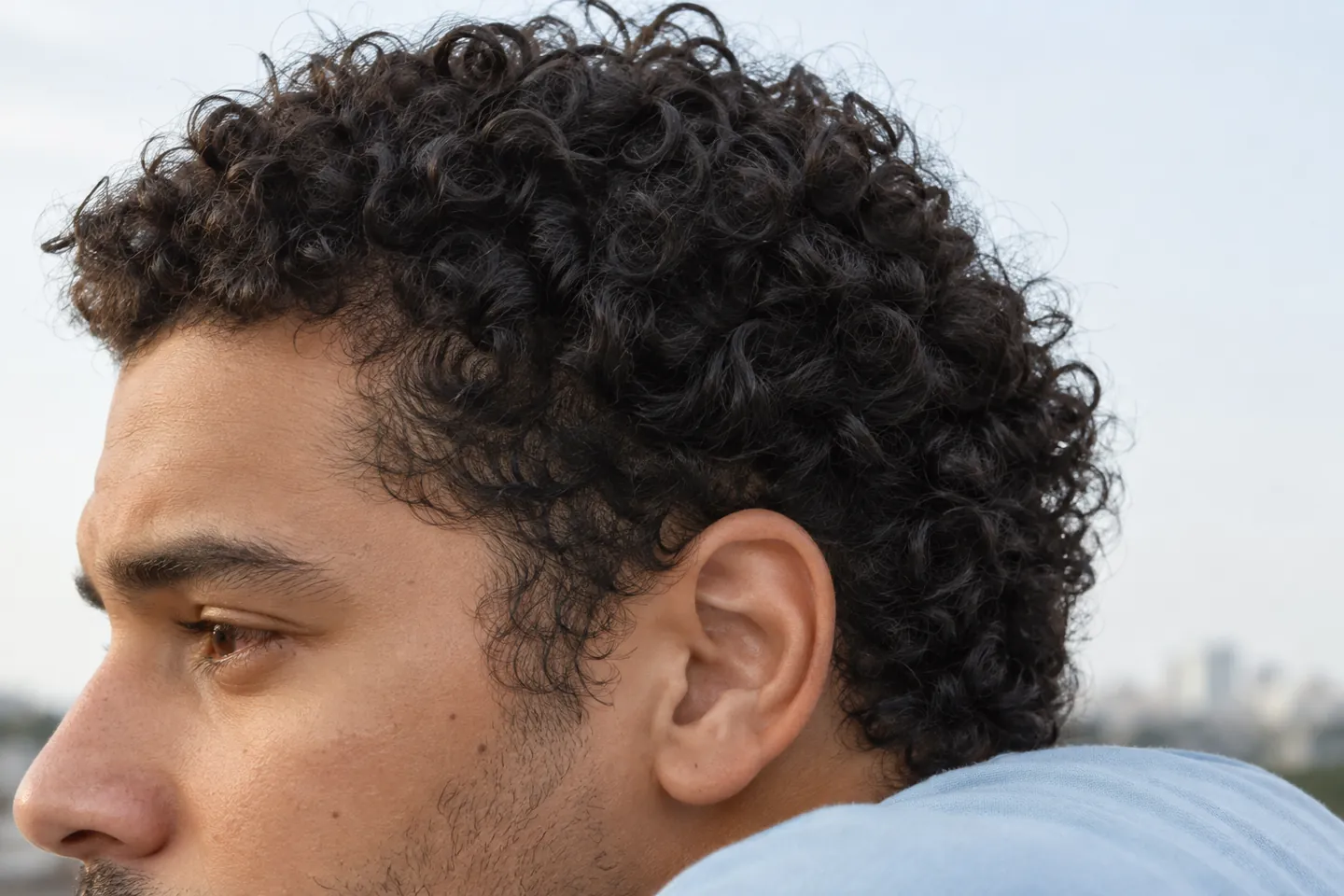 Vue rapprochée de profil montrant des boucles foncées naturelles, une implantation ordinaire à la tempe, une oreille et quelques petits cheveux devant une épaule en coton clair