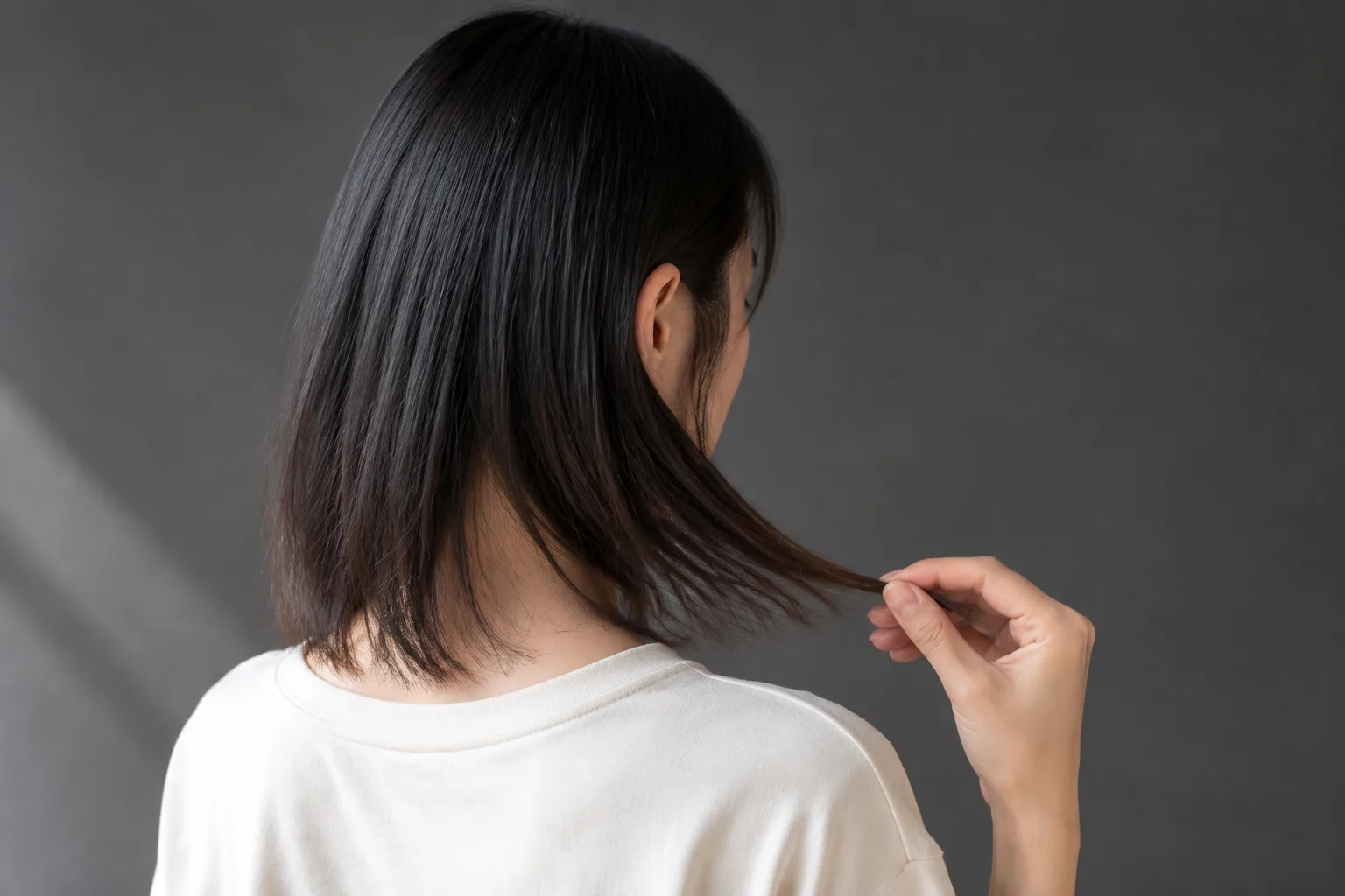 Young East Asian woman holding one side of her fine straight black hair forward against a cream shirt to inspect the lower edge