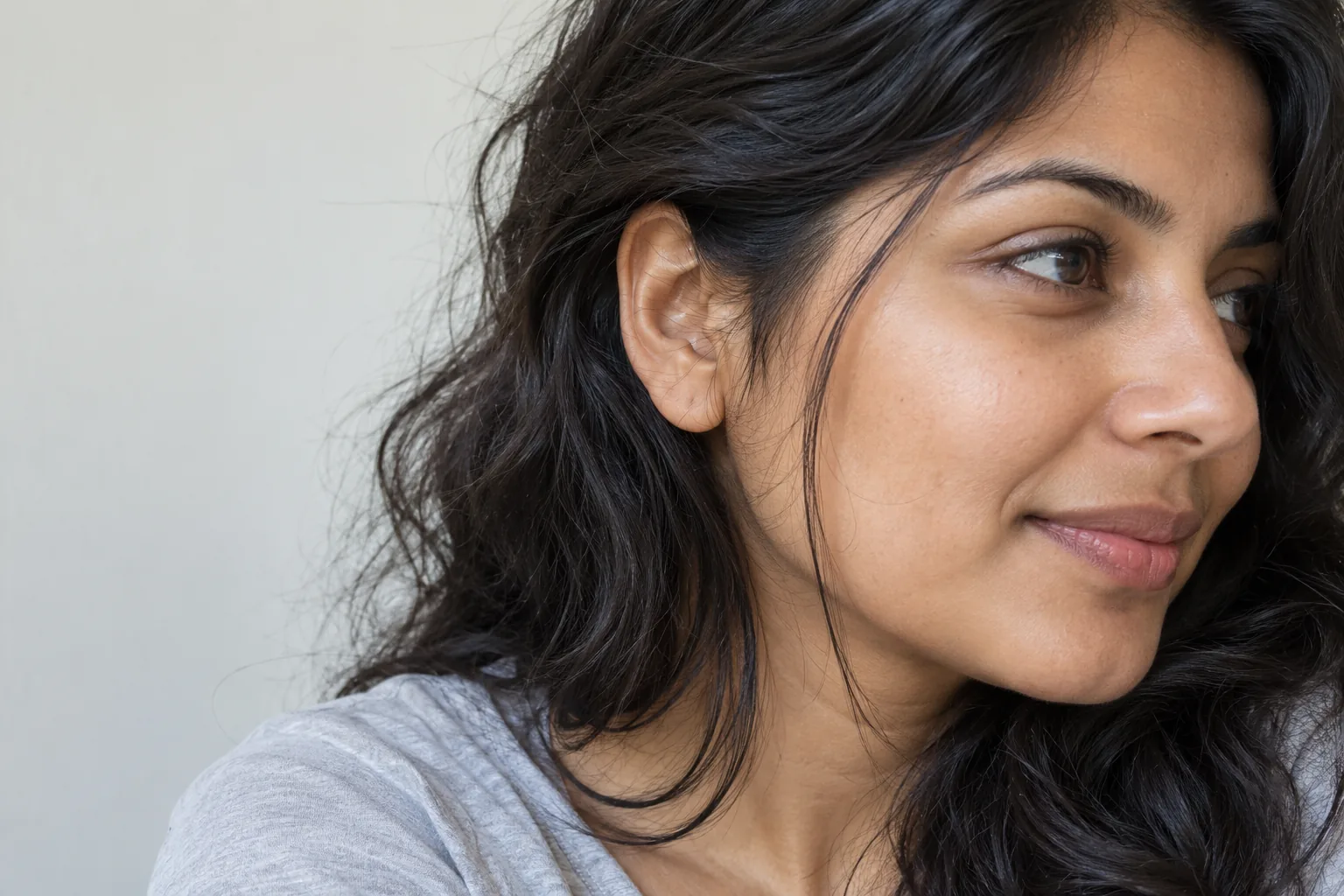 Close view of an adult’s dark wavy temple hair, fine flyaways, ear and strands resting over a light cotton shoulder