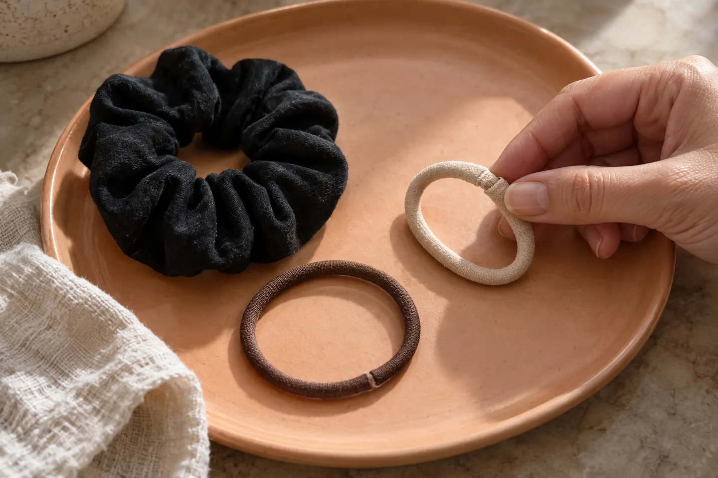Adult hand inspecting the seam of a fabric-covered hair tie beside two other plain ties on a ceramic tray