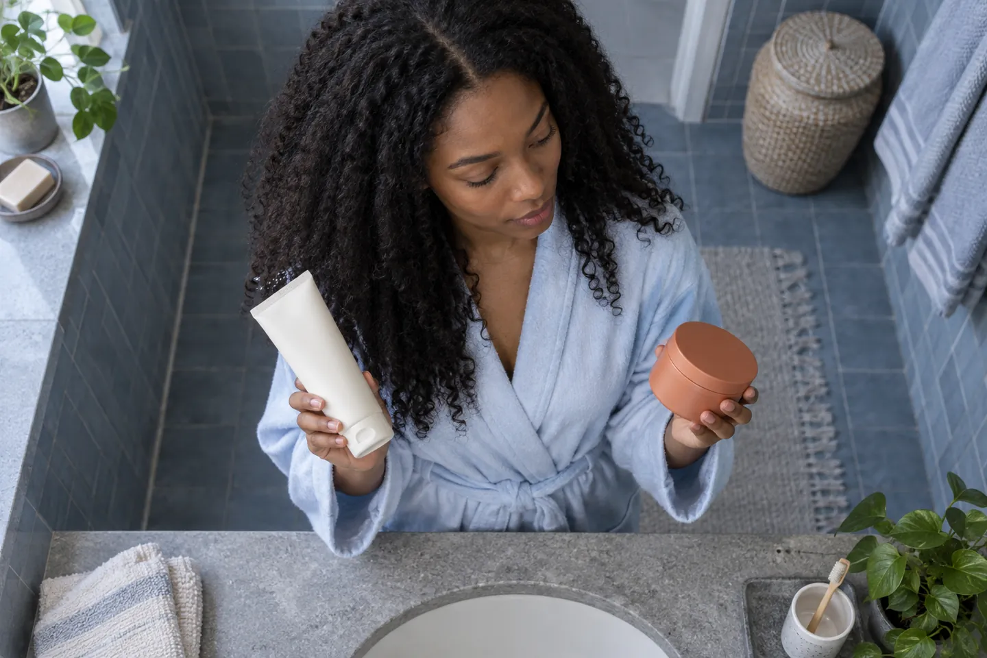 Overhead view of a young woman with long coils comparing a plain tube and a terracotta jar at a blue-grey bathroom vanity
