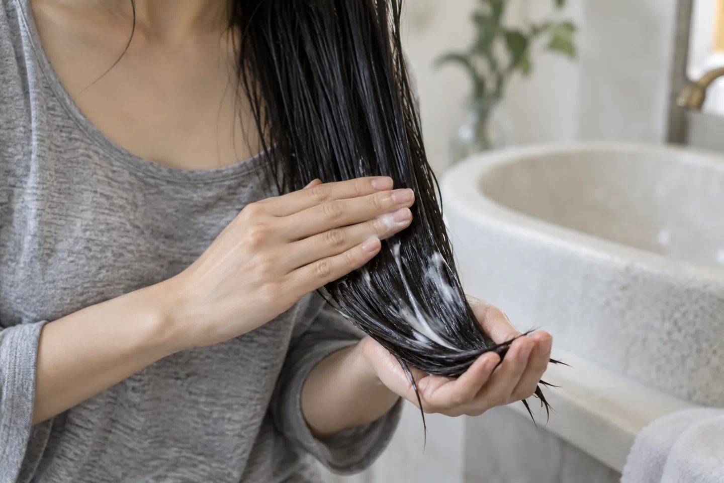 Adult hands support long damp straight hair while gently distributing a small amount of white conditioner over the lower lengths