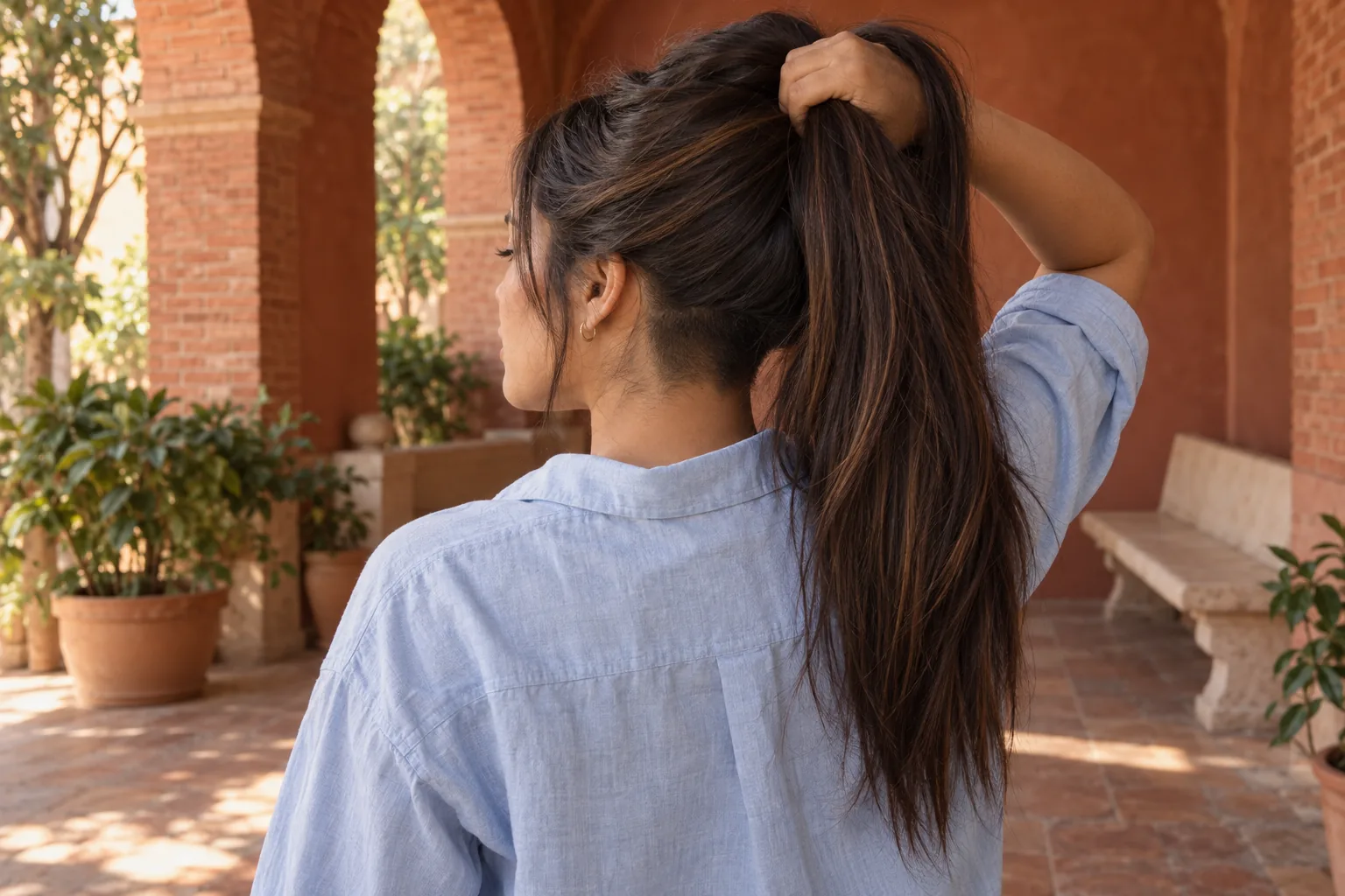 Young adult woman lifts long chestnut hair to reveal a small nape undercut in a terracotta courtyard