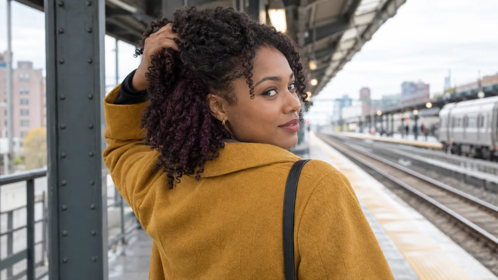Junge Frau mit Locken hebt auf einem überdachten Bahnsteig das Deckhaar und zeigt einen gedämpft pflaumenfarbenen Akzent