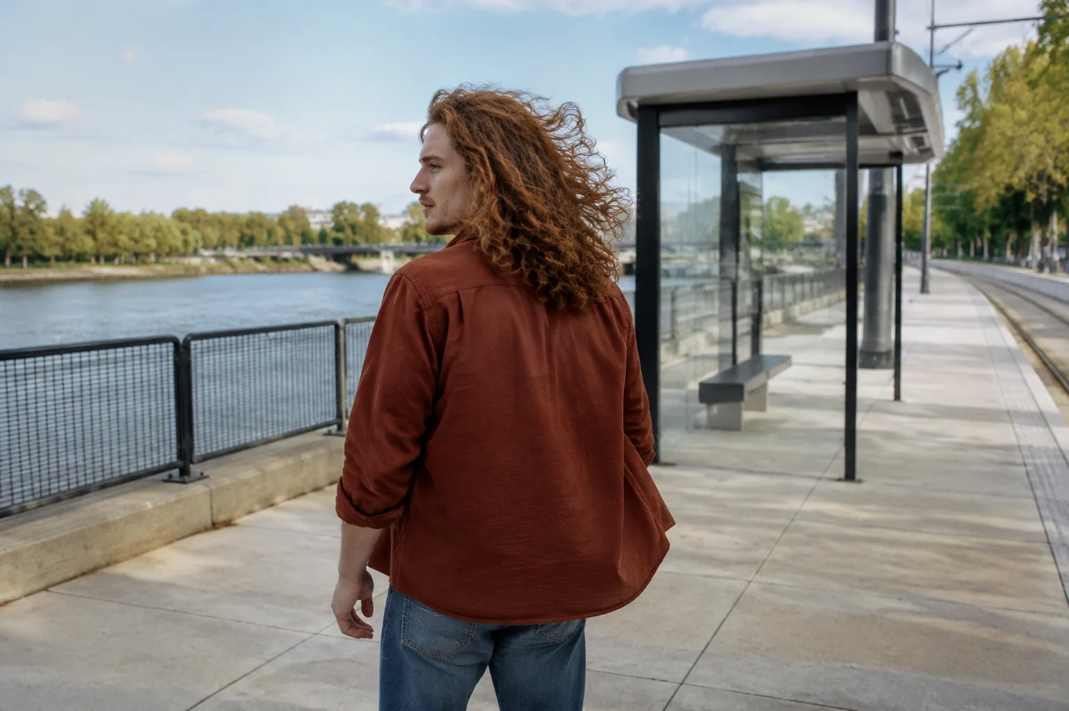 Young man with dense shoulder-length auburn waves standing at a breezy riverside tram stop to observe natural movement