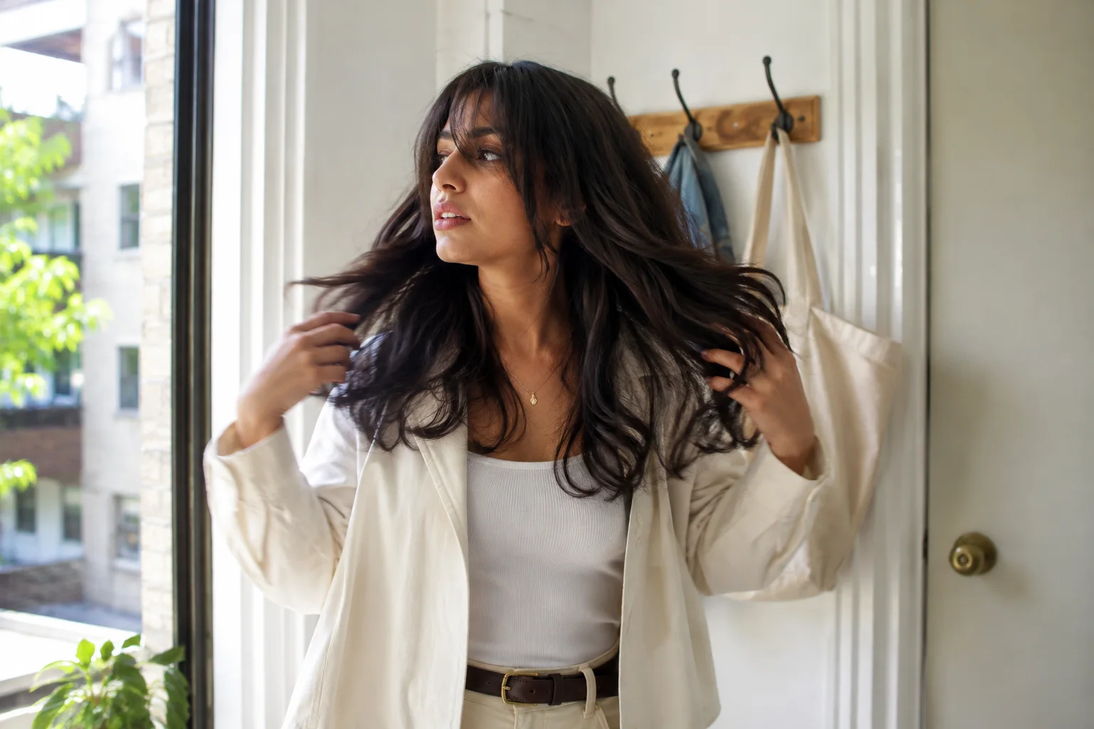 Young woman shaking out naturally air-dried layered waves beside a bright apartment window before leaving home